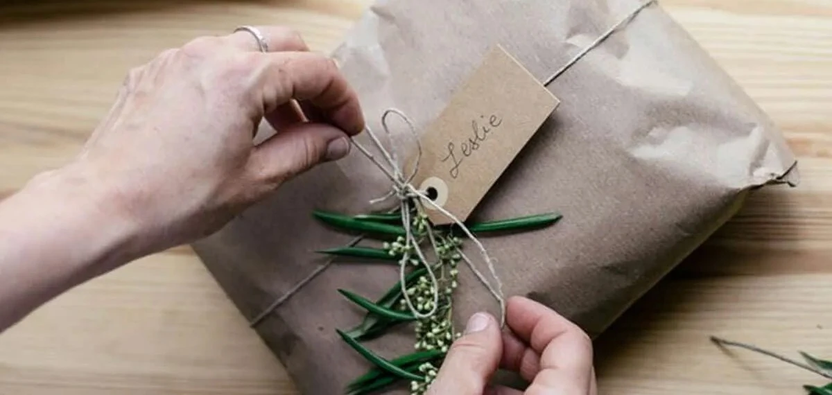Person's hands tying twine around a brown paper gift, on a wooden table.